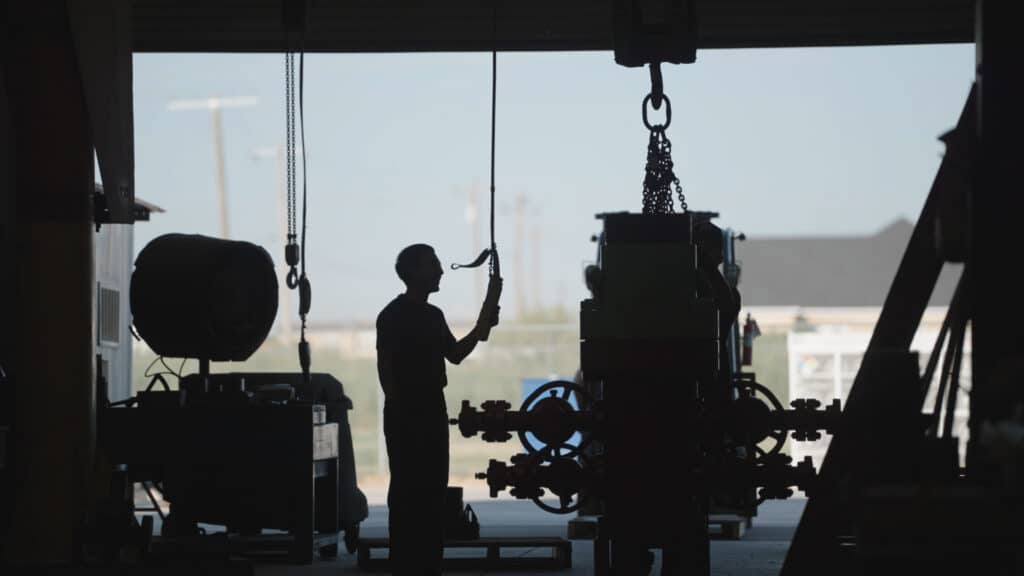 A silhouette of a worker in the Stream-flo USA shop lifting a wellhead using a crane.